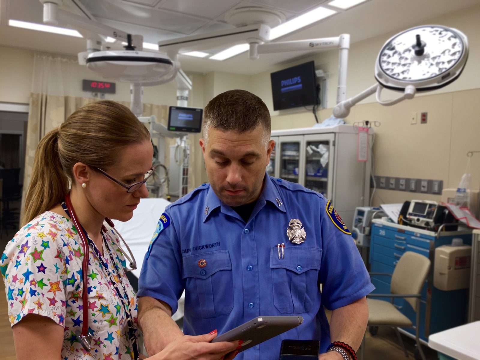Staged patient handover: Rom Duckworth, in uniform as a fire captain, reviews patient information on a tablet with a clinician in an emergency department treatment room.