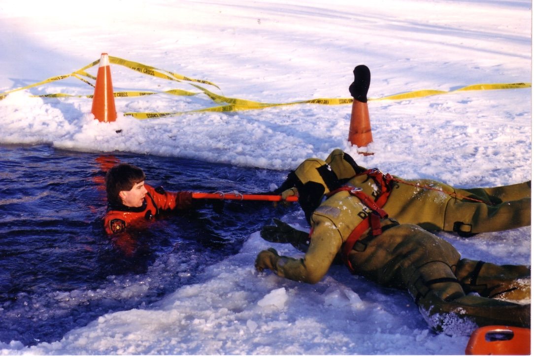An ice rescue training evolution: a rescuer on the ice reaches a person in the water with a pole.